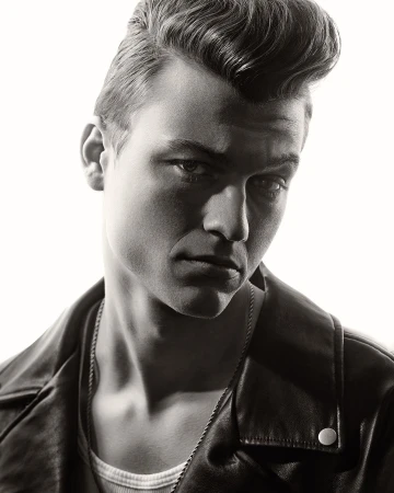 Monochrome portrait of a handsome adult male with pompadour hairstyle, wearing a leather jacket and chain necklace, against a white backdrop.